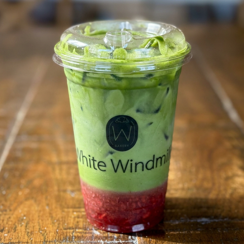 Layered green matcha drink with red fruit puree in a clear White Windmill Bakery cup on a wooden table.