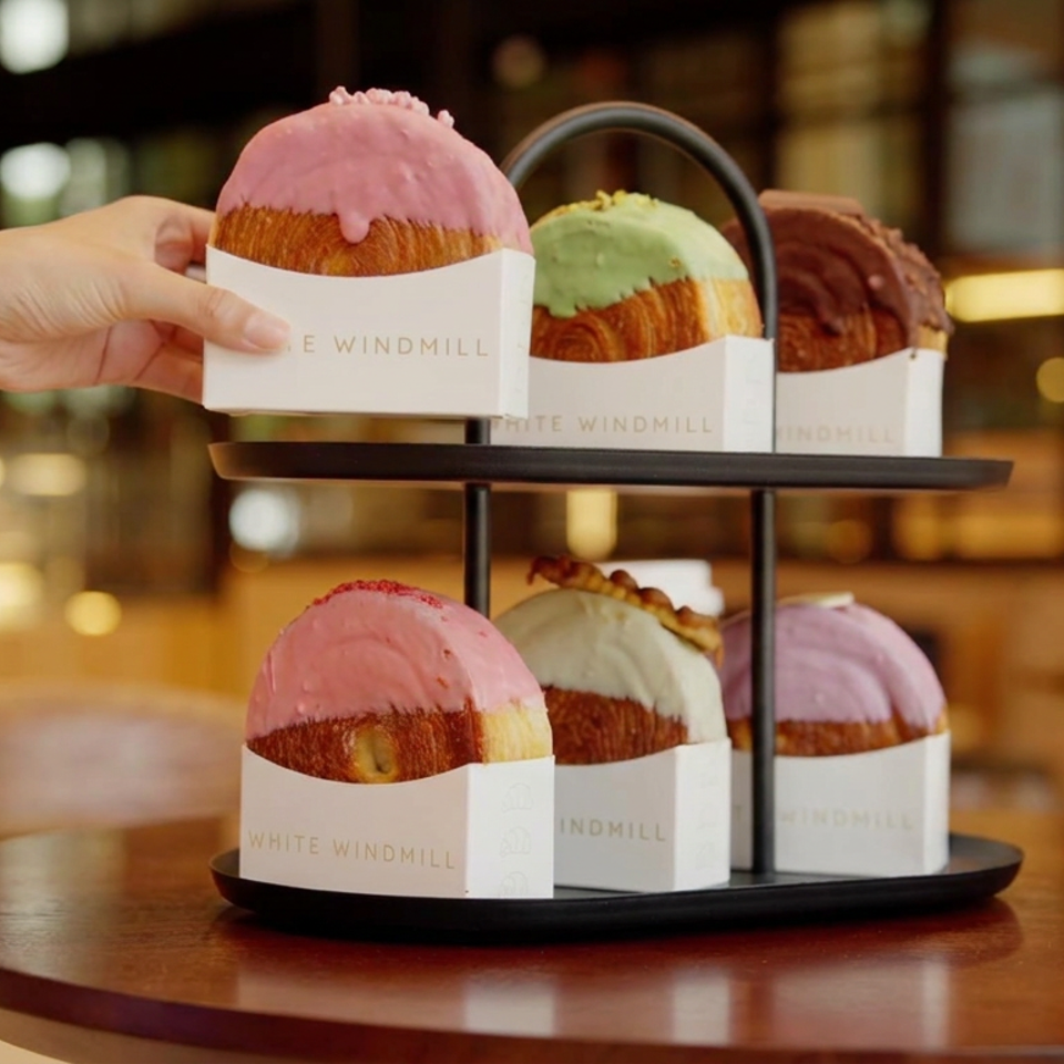 Tiered stand displays six colorful frosted pastries in White Windmill sleeves on a café table, with a hand reaching for one.