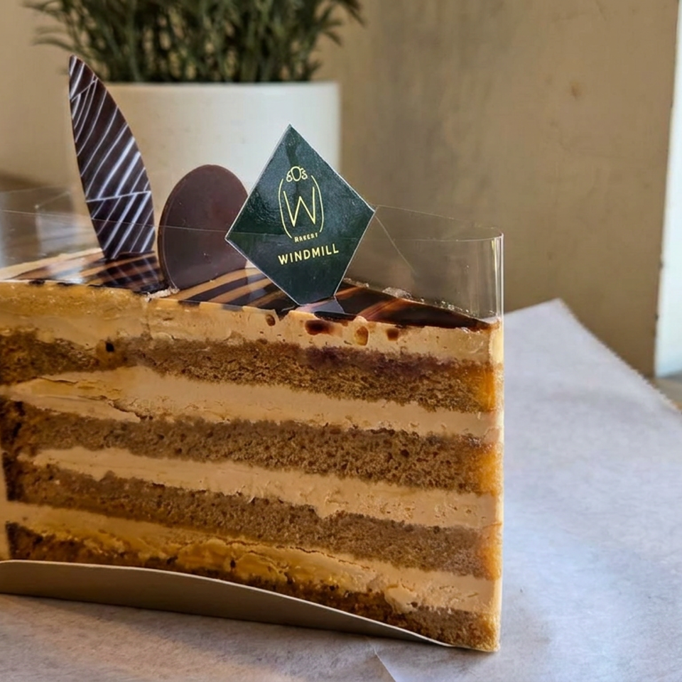 Layered coffee cake slice with cream filling and chocolate decorations sits on a café table, branded with a Windmill Bakery tag.