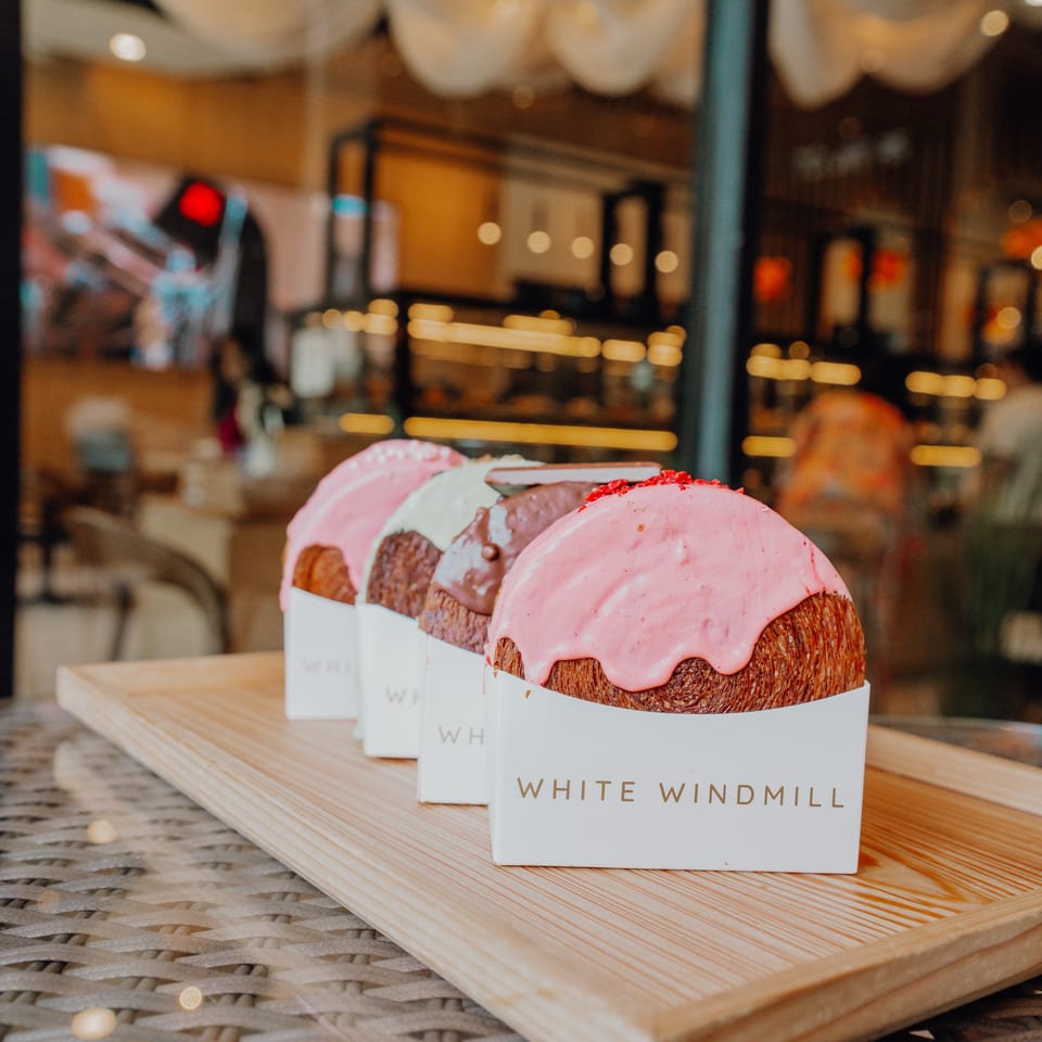 Pink-glazed pastries in White Windmill Bakery boxes sit on a wooden tray beside a softly lit café window.