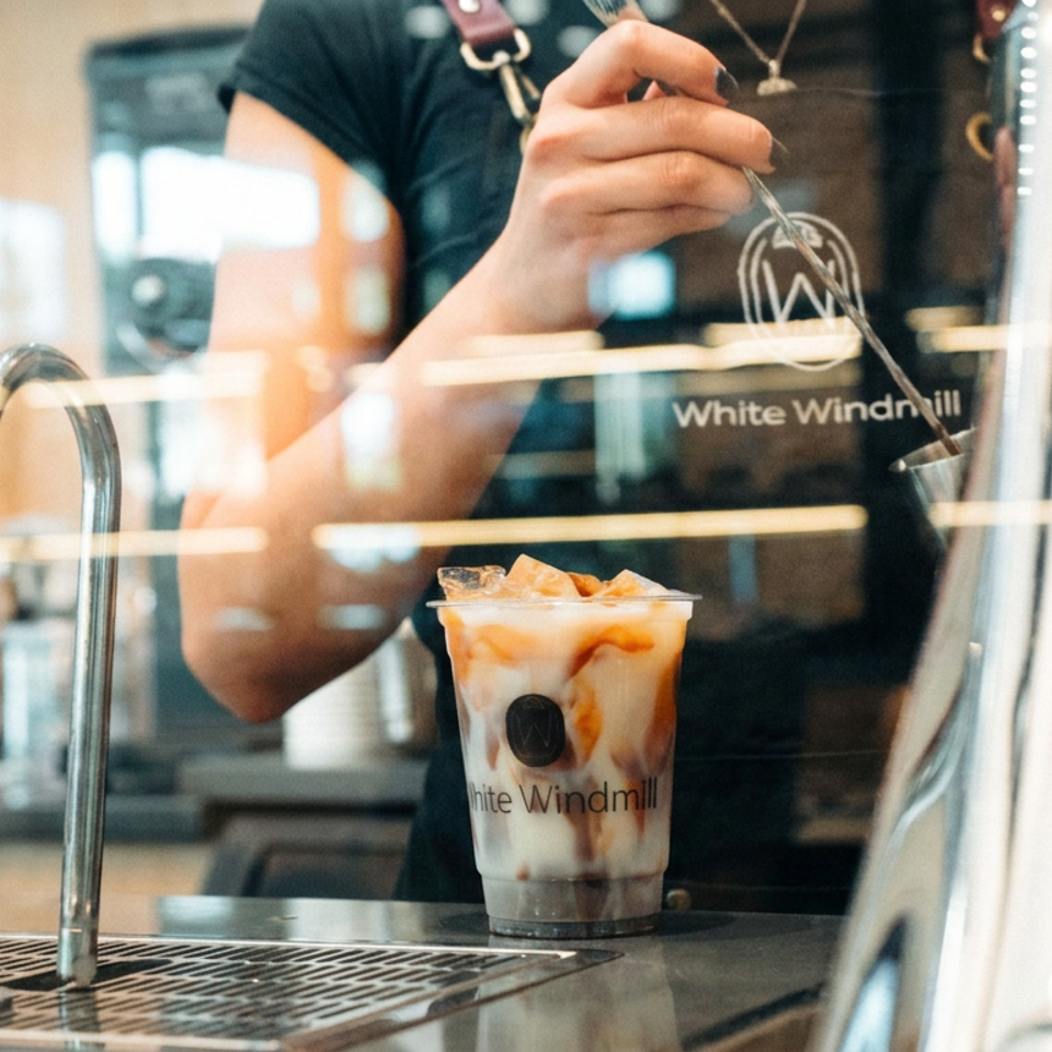 Barista prepares an iced, layered coffee drink branded White Windmill behind a café counter.