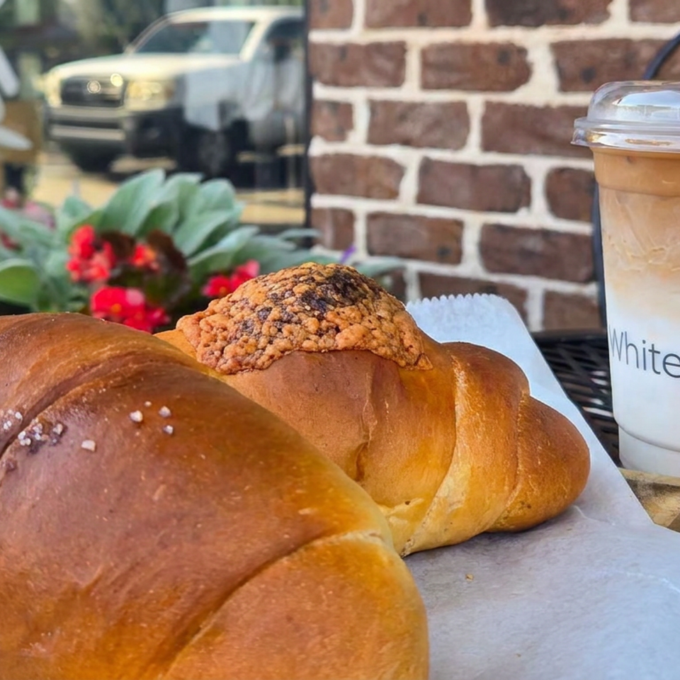 Golden braided pastries sit beside an iced coffee branded White Windmill on an outdoor café table.