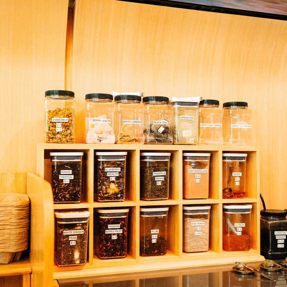Wooden shelving displays labeled jars of tea, herbs, spices, and powdered ingredients behind a restaurant counter.