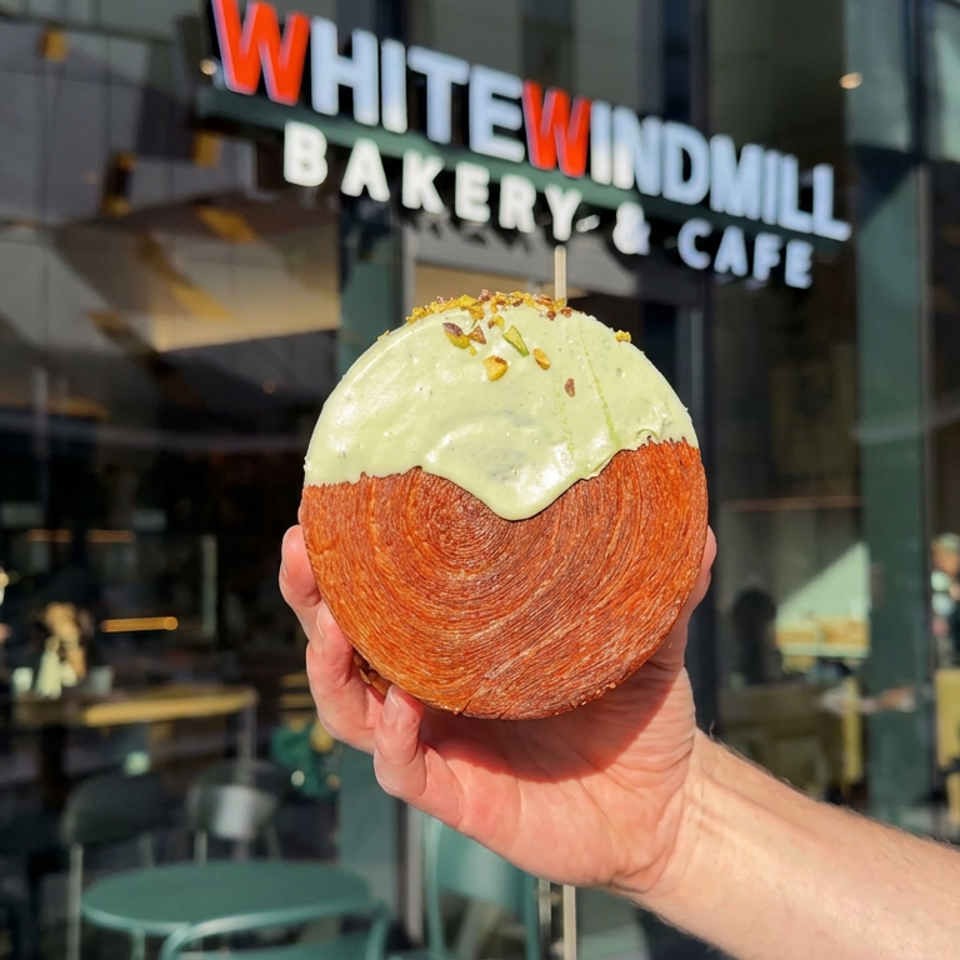 Hand holding a round pastry topped with pale green frosting and chopped nuts outside White Windmill Bakery-Cafe.